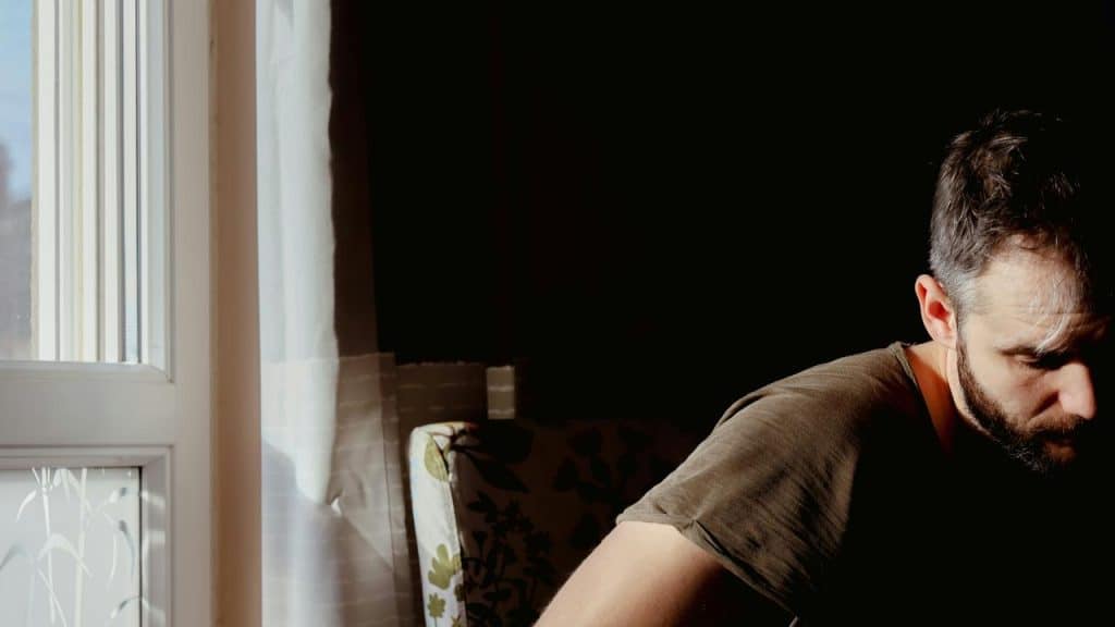 A man sitting near a window in low light, looking down pensively.