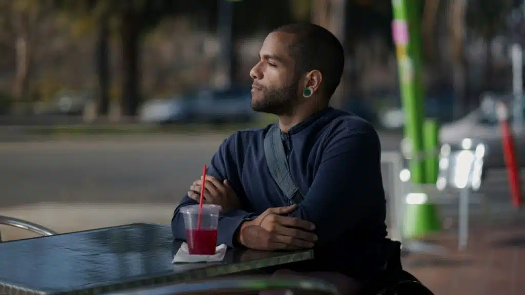A man with short hair sits at an outdoor table with a drink, looking away.