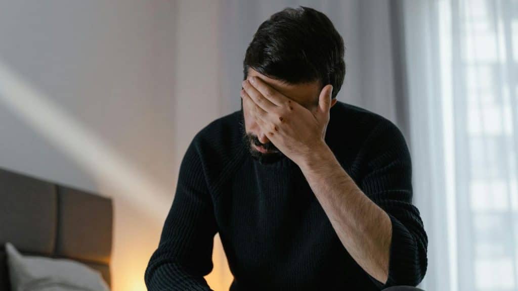 A bearded man in a dark sweater sits indoors with his hand covering his eyes and forehead.