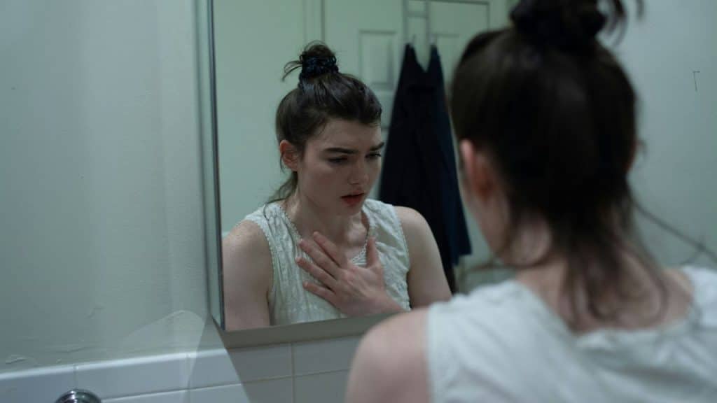 A woman with her hair in a bun looks at her reflection in a bathroom mirror.