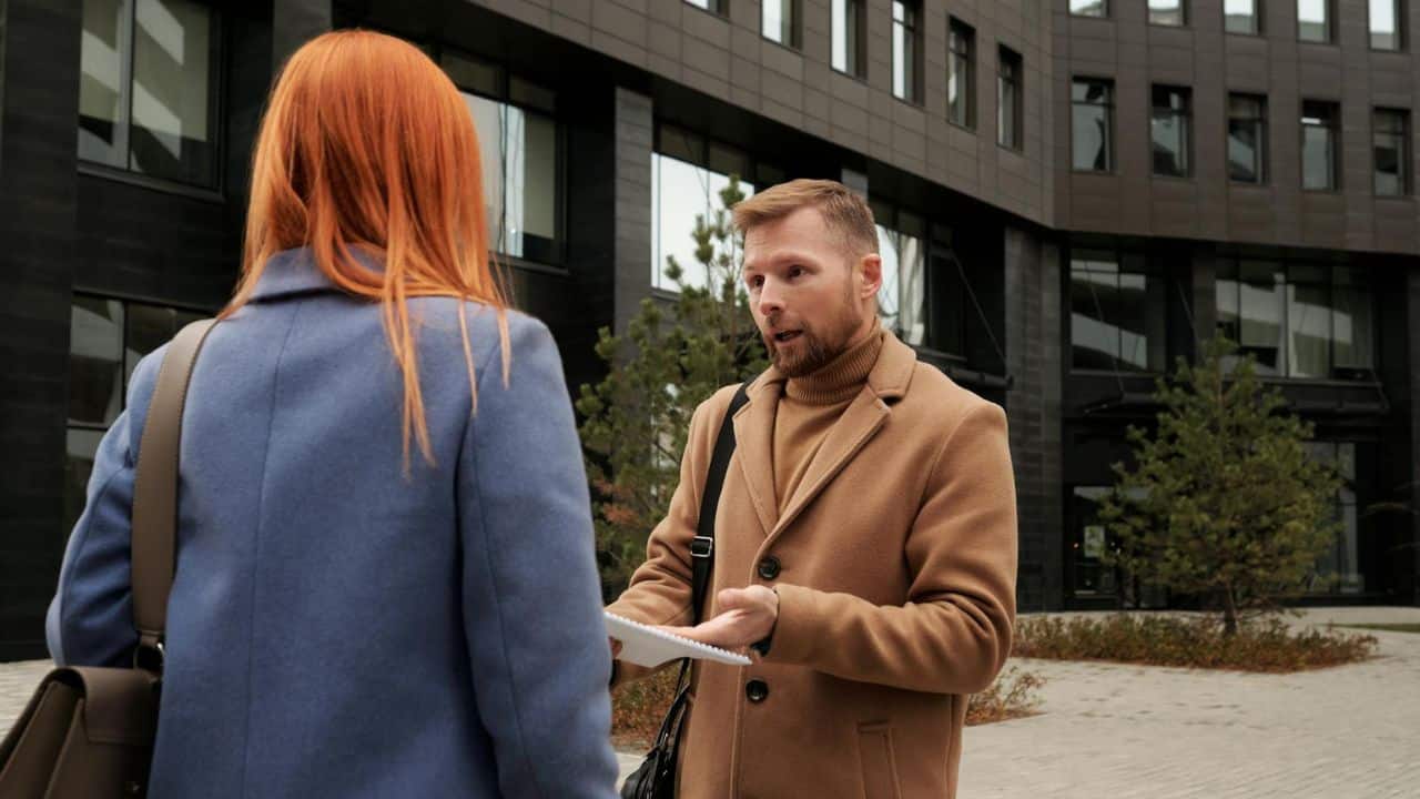 A man in a tan coat gestures while talking to a woman with long red hair.