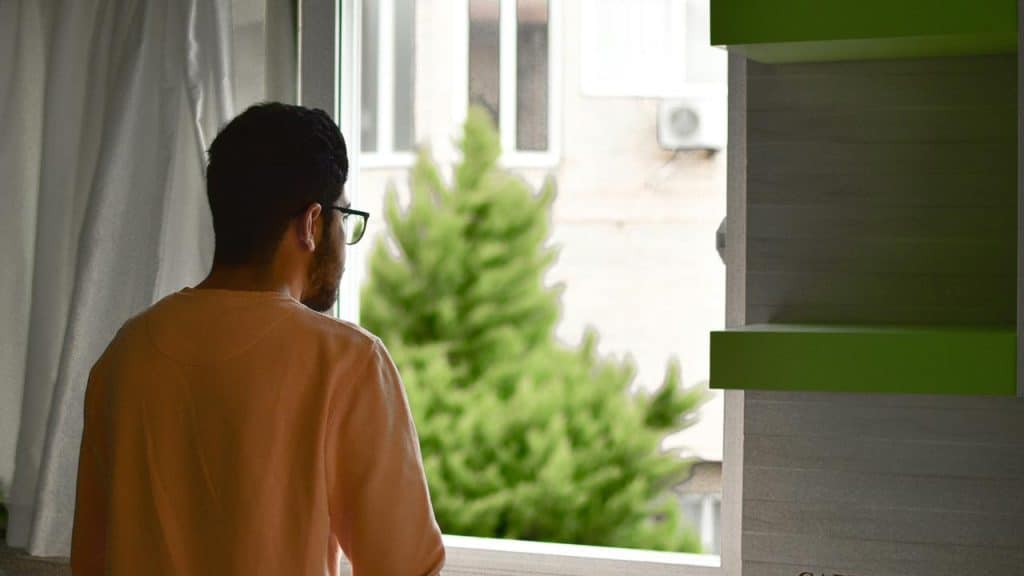 A man in glasses standing by a window and looking outside.