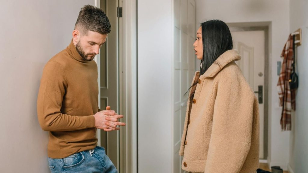 A man in a brown sweater leans against a wall while looking down at a woman.