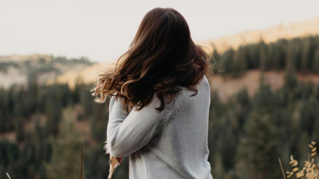 A woman standing outdoors with her back turned, looking at a forested landscape.