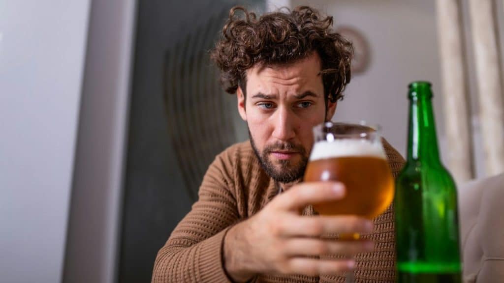 A man with curly hair looks at a glass of beer, holding it in his hand.