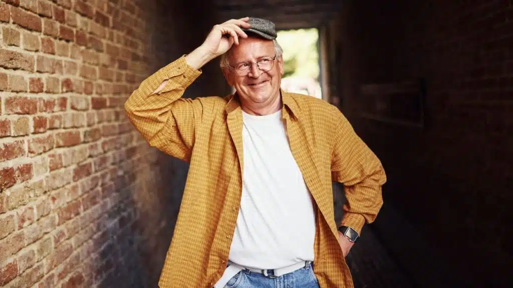 A smiling man in a yellow checkered shirt tips his flat cap in a brick alleyway.