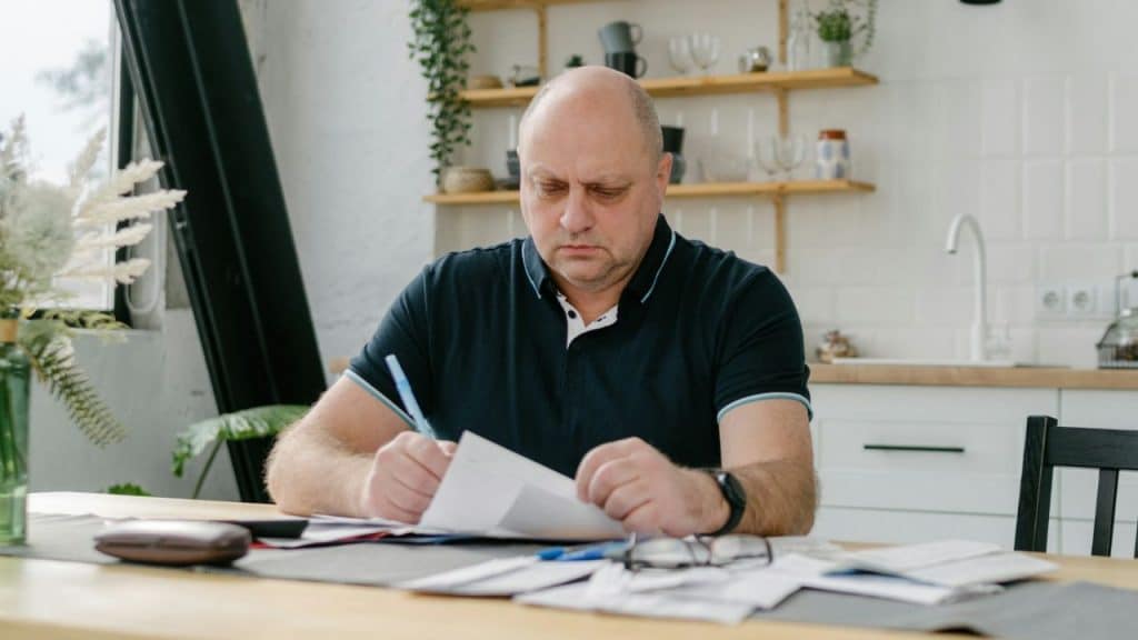 A man in a black polo shirt sits at a table reviewing and signing paperwork.