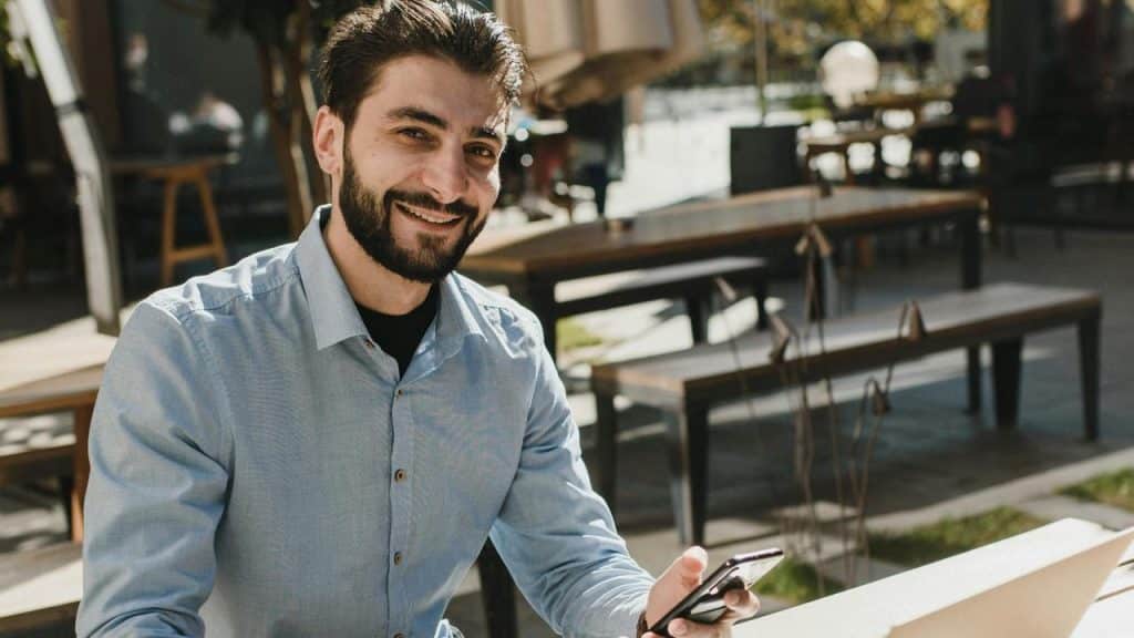 A man with a beard and blue shirt smiles while holding a smartphone outdoors.