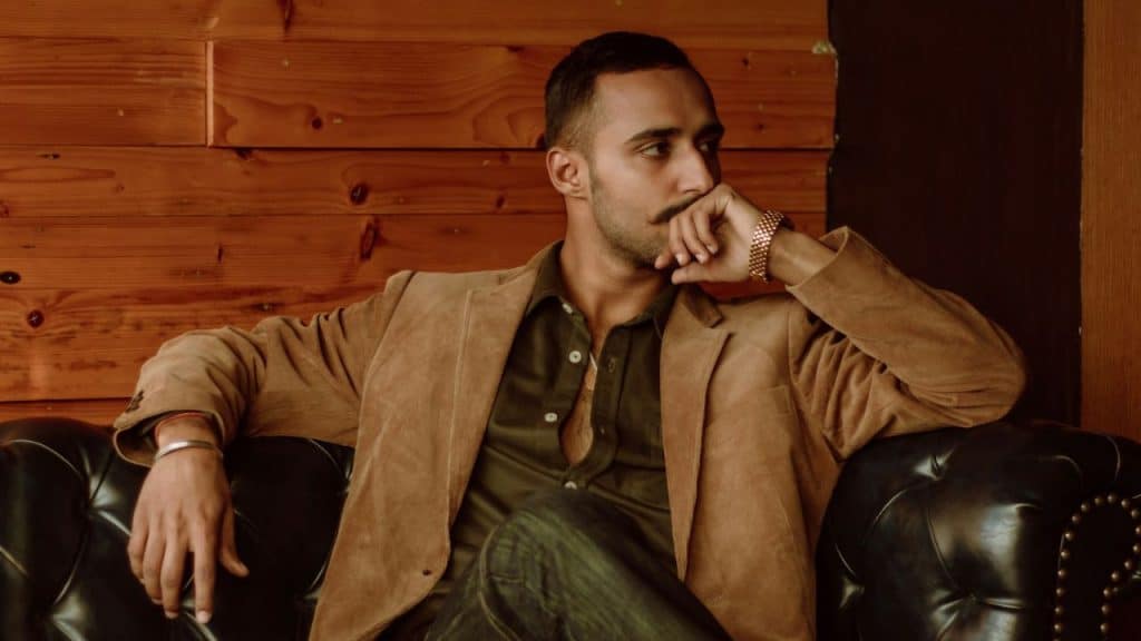 A man with a mustache in a brown jacket sits on a leather sofa against a wood-paneled wall.