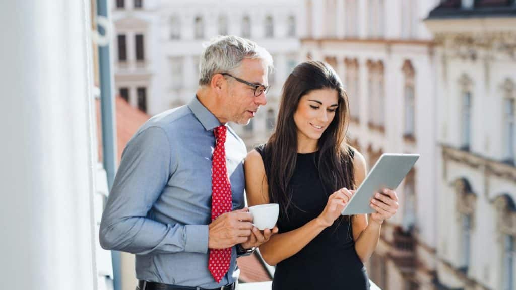 A man in a blue shirt and red tie looks at a woman's tablet outdoors.