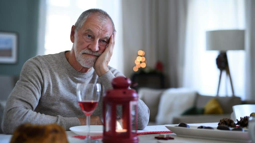 A senior man sits alone at a dining table with a somber expression.