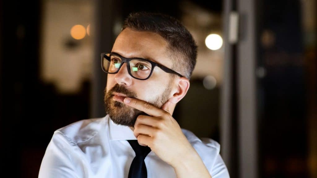 A man with glasses and a beard rests a finger on his chin while looking away.