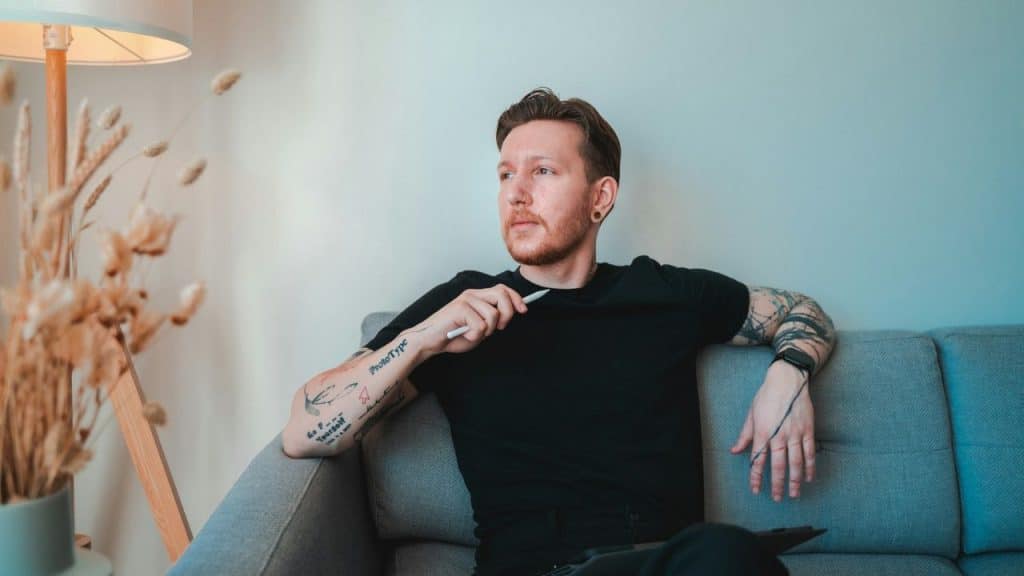 A man with tattoos is sitting on a sofa and looking to the side, holding a stylus.