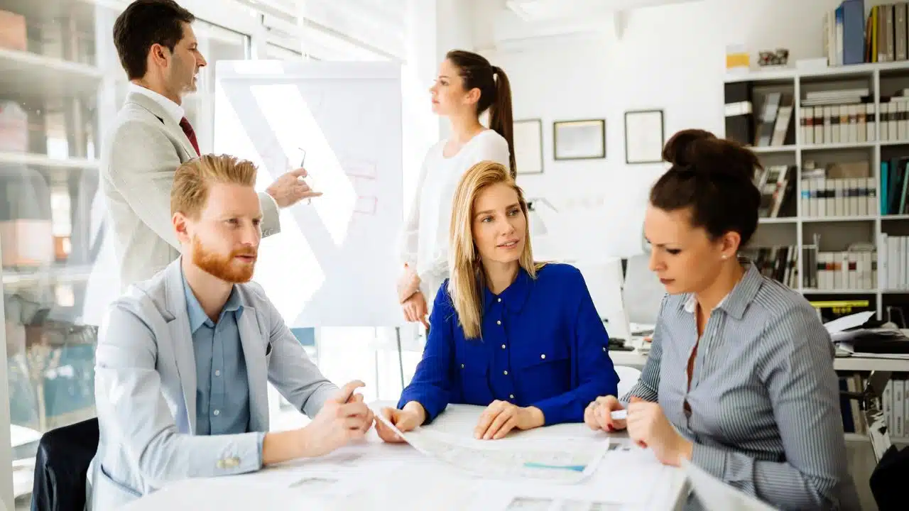 Five office colleagues in professional attire collaborate around a white table and a flipchart.