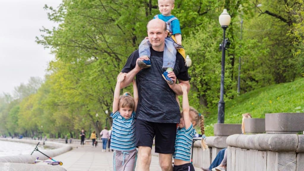 A father walking with three children, one on his shoulders and two holding his hands.