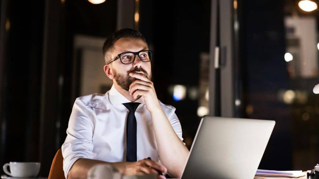 A man in an office thinking while using a laptop.