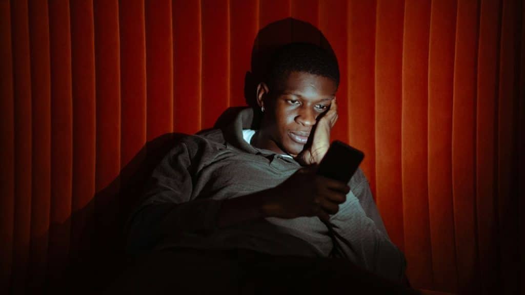 A man leans against a red pleated wall while looking down at a glowing smartphone.