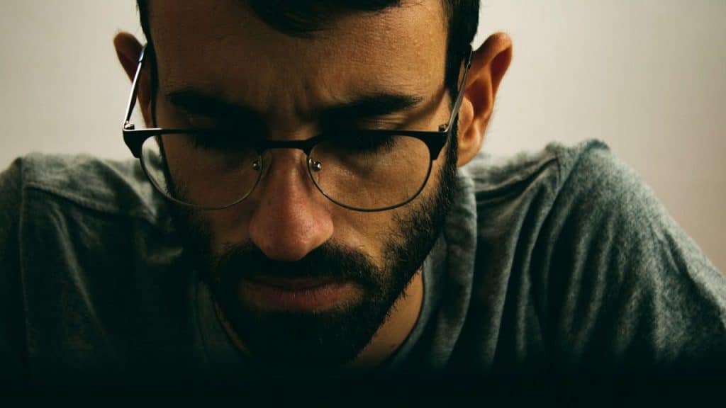 A man with a beard and glasses looks downward while wearing a grey shirt.