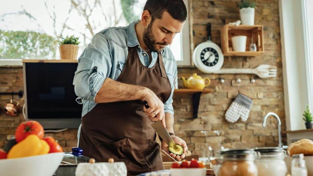 A man in an apron slicing an avocado in a kitchen.