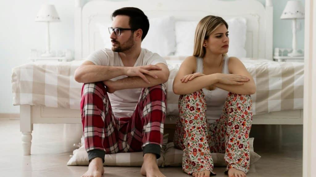 A man and a woman in pajamas sit on the floor leaning against a bed.