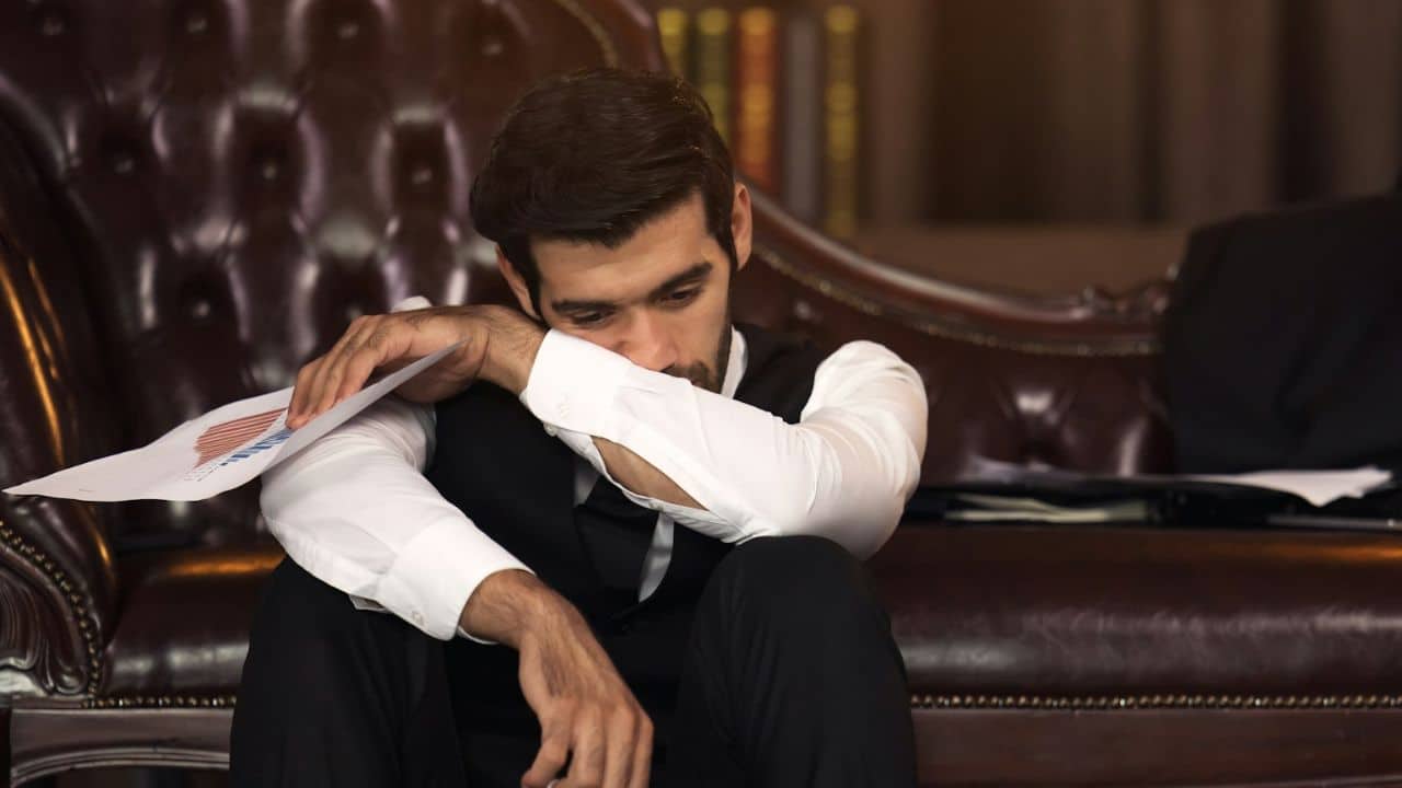 A man in a vest and white shirt sits on the floor, resting his head on his arm.