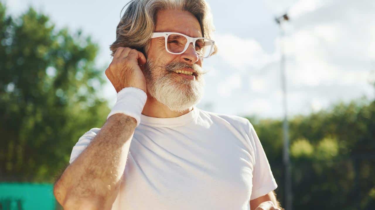 An older man with white glasses adjusting his ear outdoors.