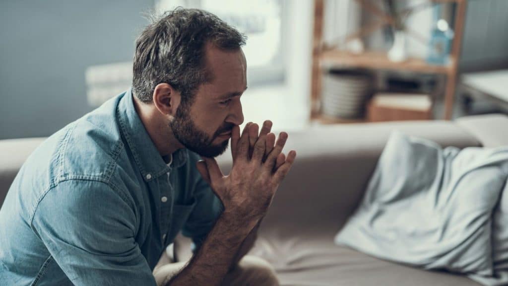 A bearded man in a denim shirt sits on a sofa with his hands clasped.