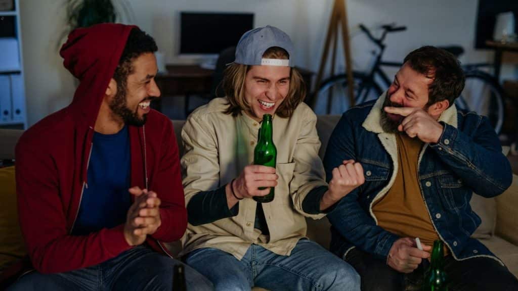Three men sit on a couch, laughing and holding green glass bottles in a room.