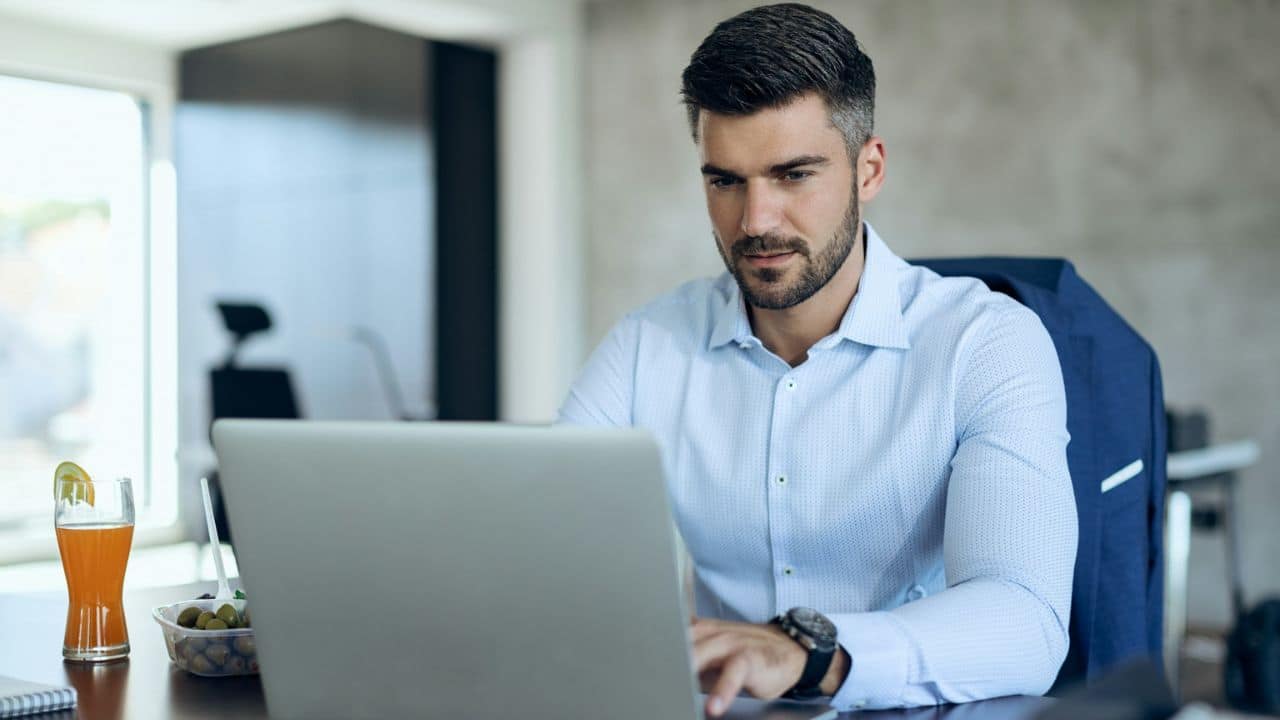 A man in a light blue shirt works on a laptop at a desk with a drink nearby.