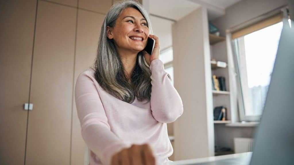 A smiling woman with long grey hair talks on a smartphone while sitting at a desk.