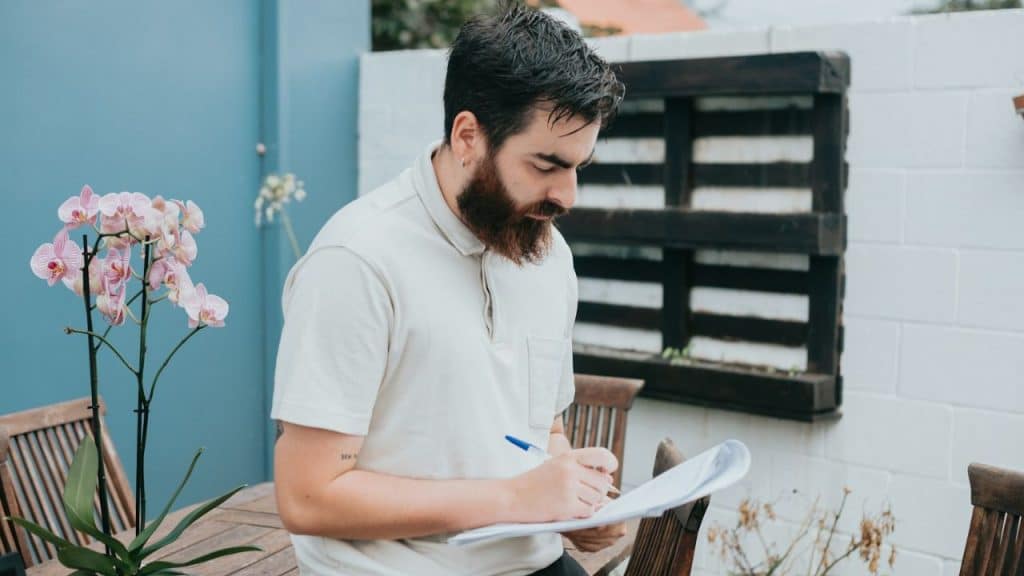 A bearded man in a polo shirt writing on a document outdoors near flowers.