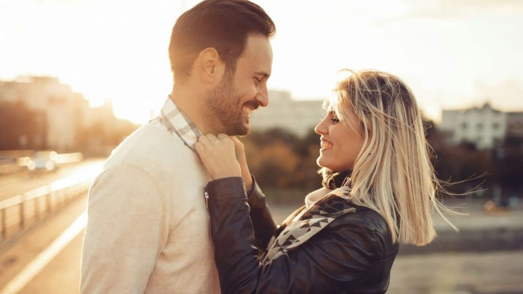 A man and woman smile at each other outdoors as she adjusts his shirt collar.