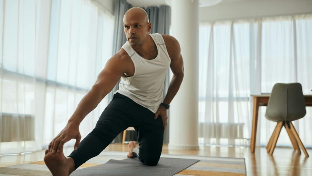 A muscular man in a white tank top stretches his leg on a mat inside a room.