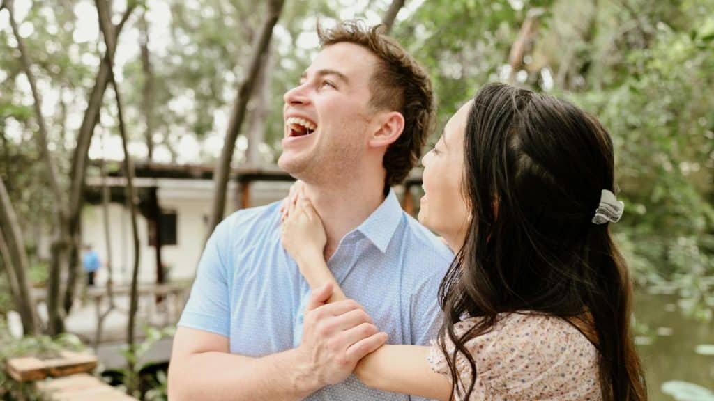 A smiling couple outdoors, laughing together among trees near the water.