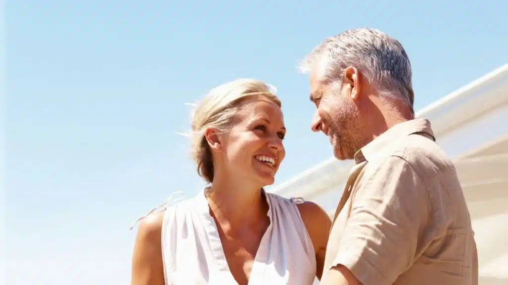 A smiling, middle-aged couple looks happily at each other outdoors under a bright sky.