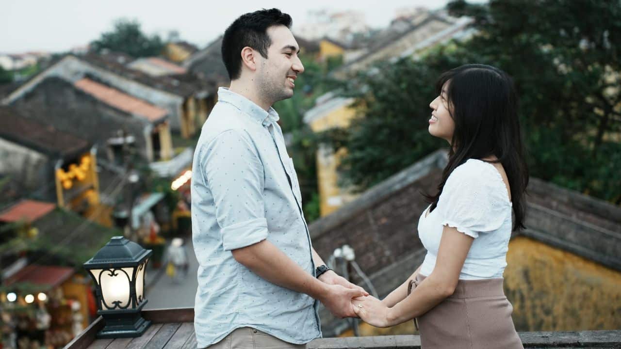 A man and woman stand on a balcony overlooking a town, smiling and holding hands.