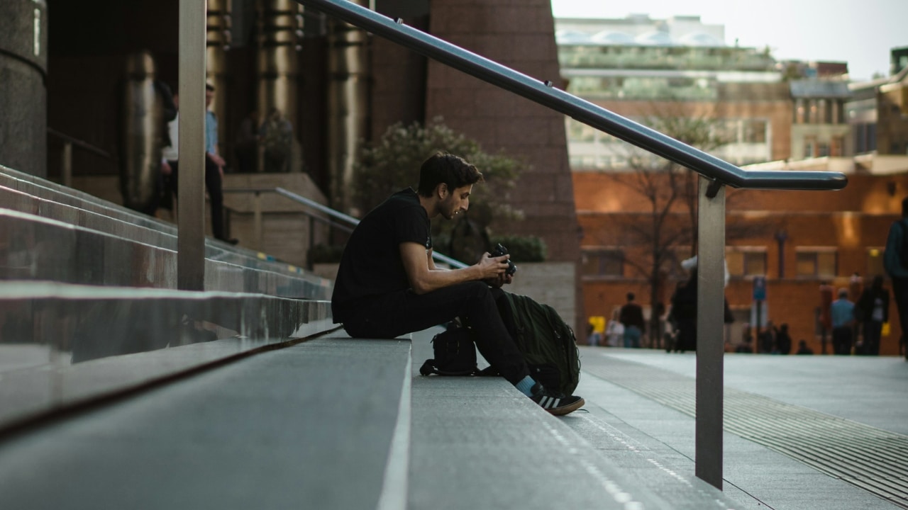 A man in a black shirt sits on concrete stairs outdoors, holding a camera.
