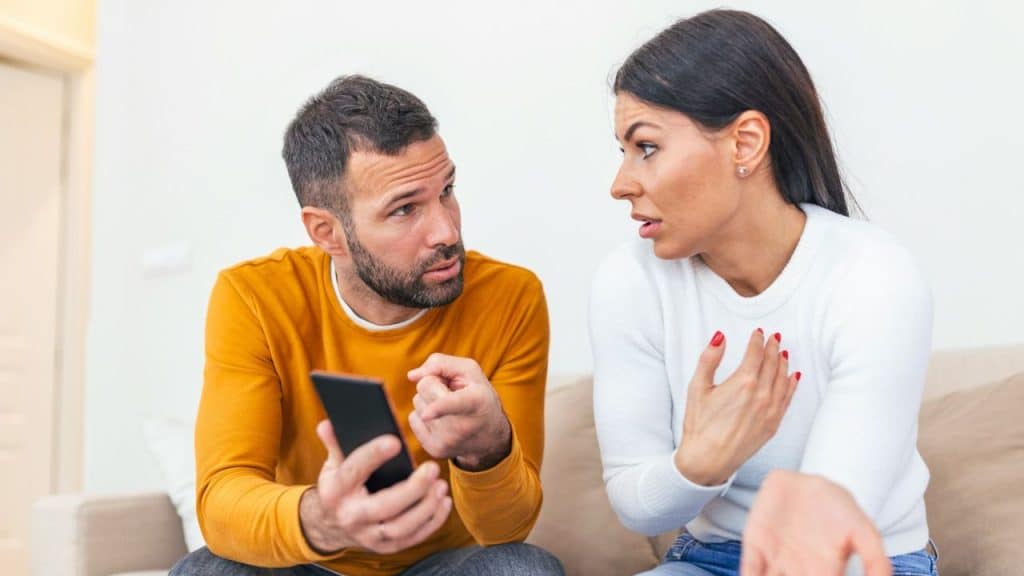 A man holds a smartphone while looking at a woman who is gesturing and speaking.