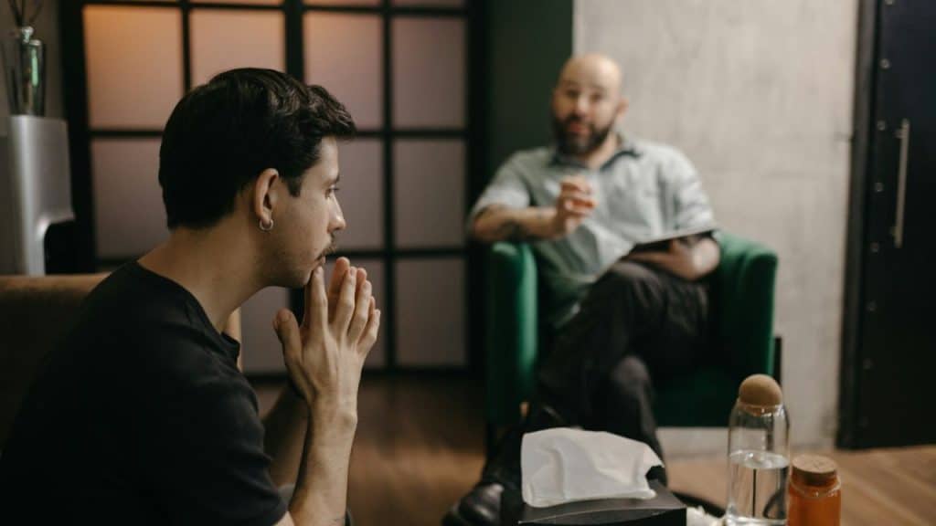 A man sits with his hands clasped to his face, listening to another man speaking.