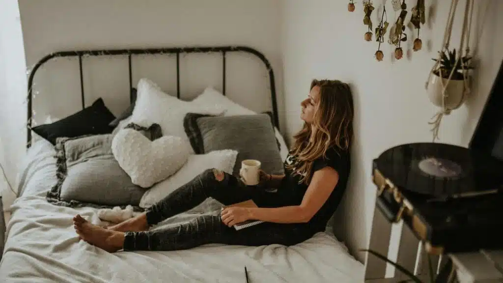 A woman sitting on a bed holding a mug and looking thoughtful.