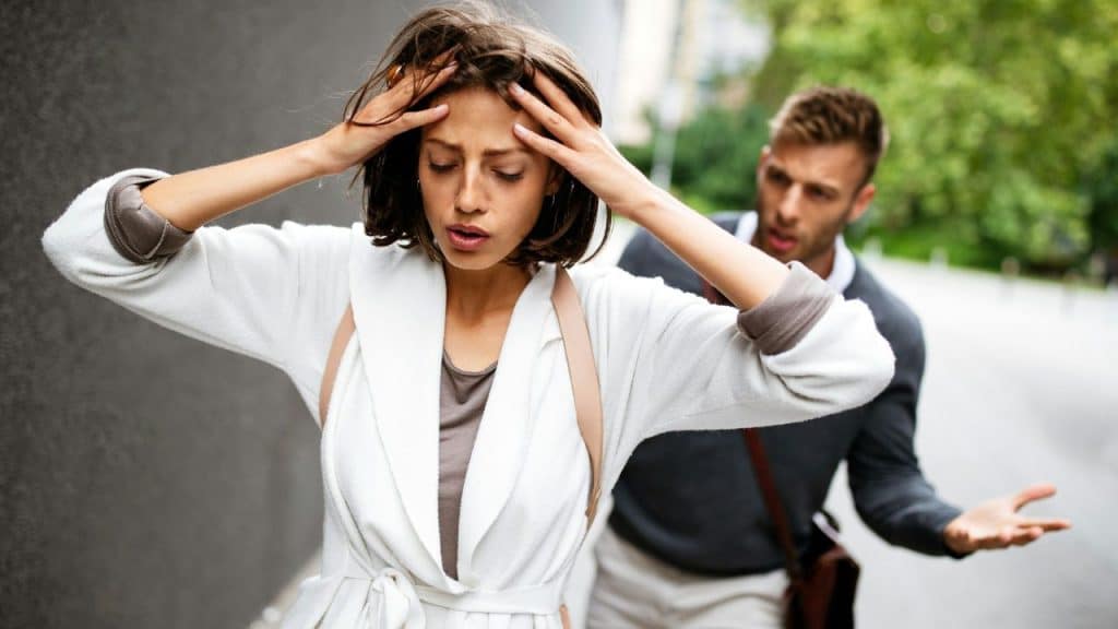 A woman holds her head while a man in the background gestures with open hands.