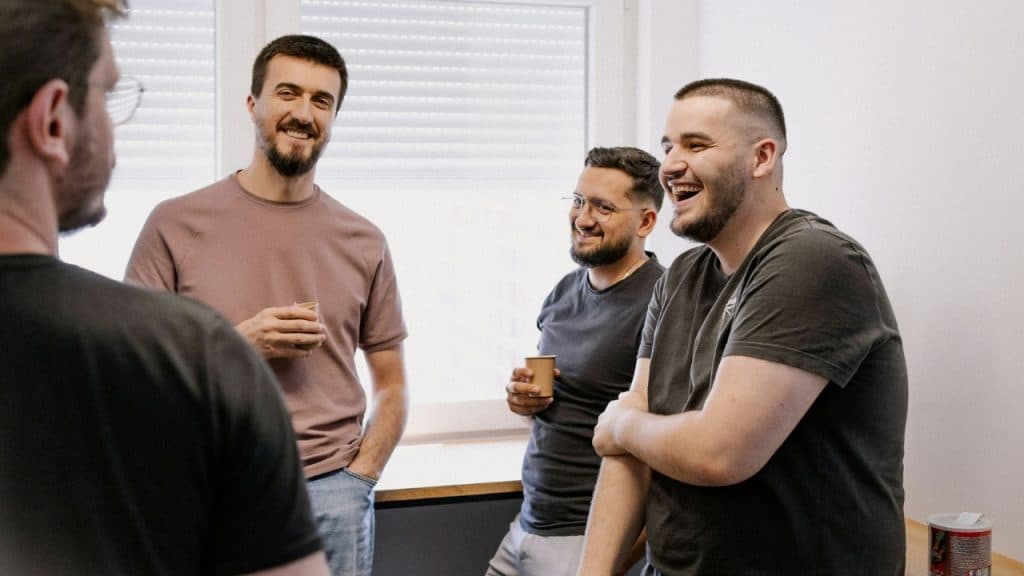 Four men stand together in a room, smiling and laughing while holding small paper cups.