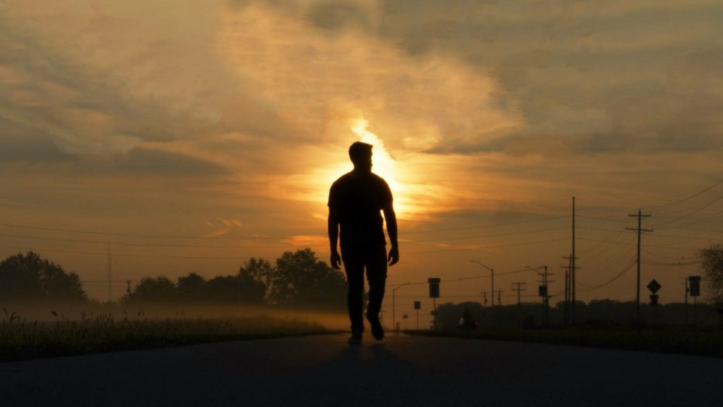 A silhouette of a person walks down an empty road toward a glowing sunset.