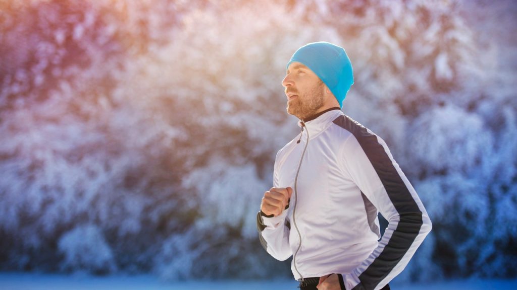 A man in a blue beanie and athletic jacket runs through a snowy landscape.
