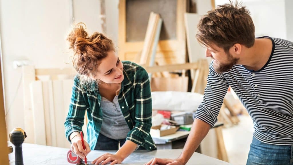 A man watches a woman mark a piece of wood with a pencil and tape measure.