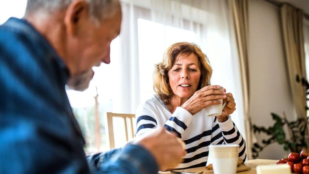 A senior woman holds a teacup, looking at a man sitting across from her.