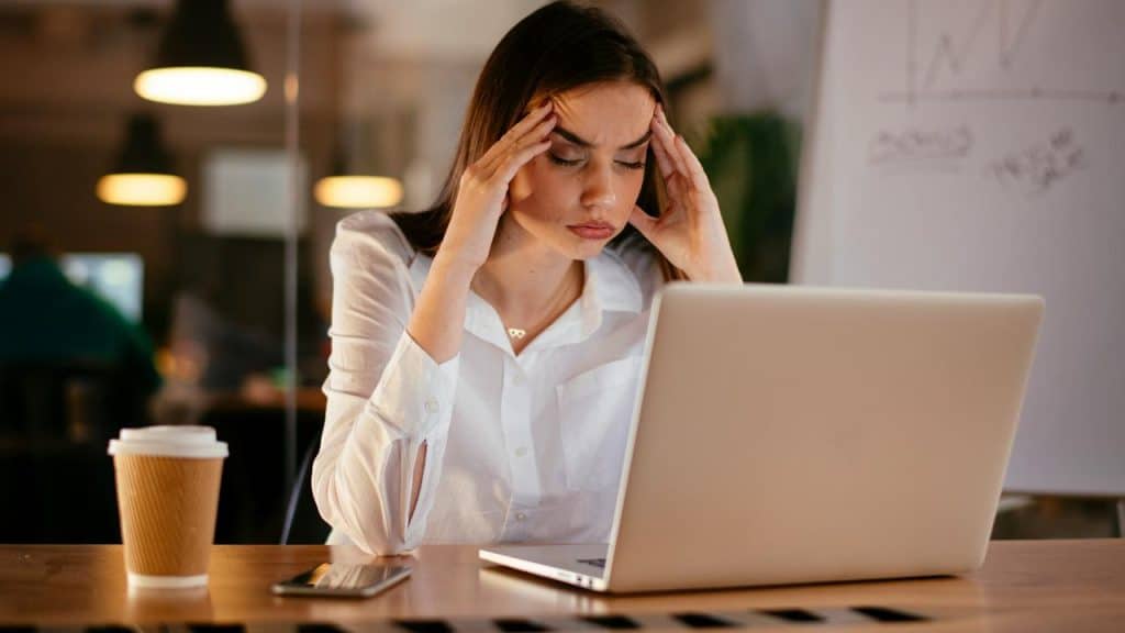 A woman in a white shirt looks stressed while working on a laptop at a desk.