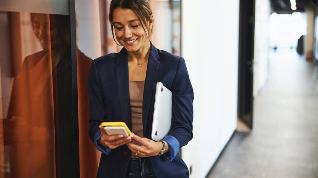 A woman in a blazer smiling at her phone while holding a laptop.