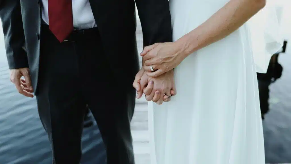 A bride in a white dress and a groom in a dark suit holding hands on a dock over water.