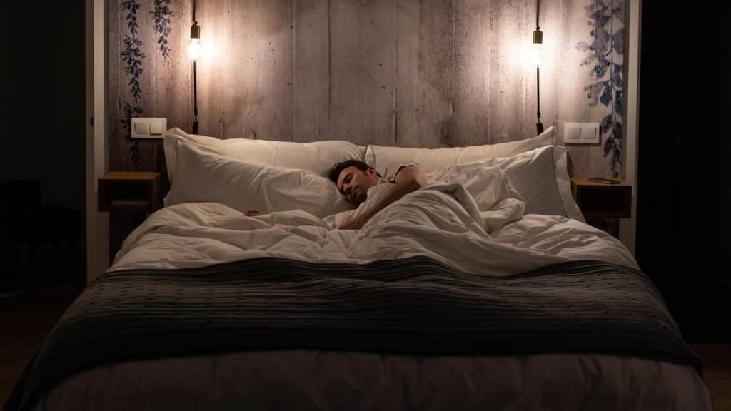 A person sleeping alone in a neatly made bed under soft bedside lights.